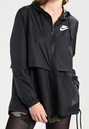 Nainen pukeutuneena mustaan Nike-huppariin, jossa on vetoketju, joustavat hihansuut ja maastokuvioiset leggingsit, käsi lähellä taskua.