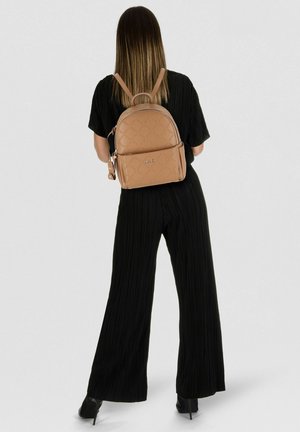 Vrouw met recht bruin haar, gekleed in een zwart geplooid outfit en met een beige rugzak, staand tegen een effen lichte achtergrond.