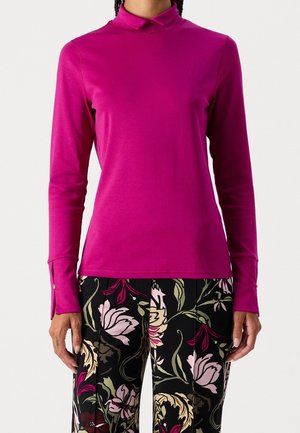 Person iført en langærmet, højhalset magentafarvet top kombineret med sorte bukser med grønne, pink og cremefarvede blomsterprints.