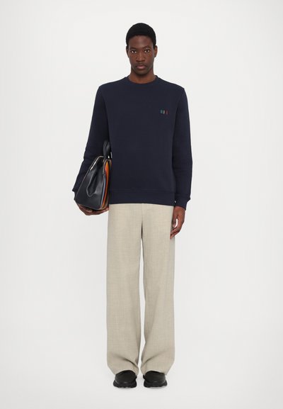 Sweatshirt marine avec de petites rayures multicolores sur la poitrine, associé à un pantalon large beige clair et des chaussures noires. Sac fourre-tout tenu à la main.