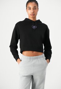 Felpa nera corta con cappuccio che presenta una tasca frontale e lacci, con un logo viola sul petto. Abbinata a pantaloni della tuta grigi chiari.