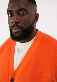 Oranges Strickcardigan mit V-Ausschnitt und einem Knopfverschluss, über einem weißen Hemd getragen. Verfügt über ein strukturiertes Gewebe und Logo-Detail.