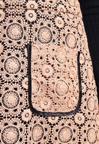 Krajková zástěra v jemně růžové barvě s intricátními květinovými vzory, se černým lemem a náprsní kapsou. Texturovaný háčkovaný design.