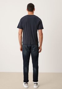 Camiseta de manga corta gris oscuro con cuello redondo, combinada con jeans oscuros de corte Slim y zapatillas blancas, exhibida desde la parte trasera.