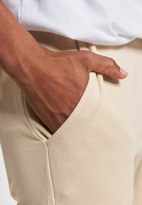 Beżowe bawełniane spodnie mają kieszeń po boku, w której spoczywa ręka. Materiał ma gładką teksturę i dopasowany krój.
