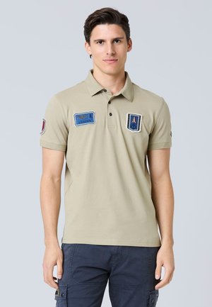 Polo di colore beige con toppe blu e rosse a contrasto sul petto e sulla manica, colletto con tre bottoni, maniche corte e una texture morbida.