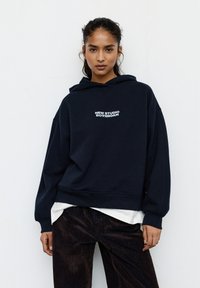 Granatowa bluza z kapturem wykonana z miękkiego materiału, z przodem ozdobionym białym napisem "NEW STUDIO ROTTERDAM", o luźnym kroju.