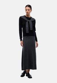 Pull noir à manches longues avec un détail de cravate grise au col, associé à une longue jupe tricotée gris foncé. Le mannequin porte des chaussures noires.