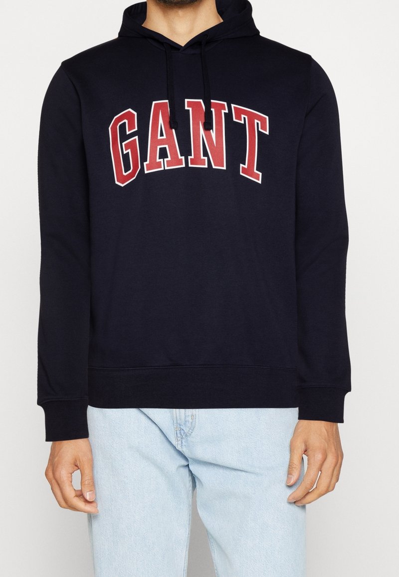 Homme portant un sweat à capuche noir avec un logo "GANT" rouge et blanc et un jean bleu clair sur un fond uni.