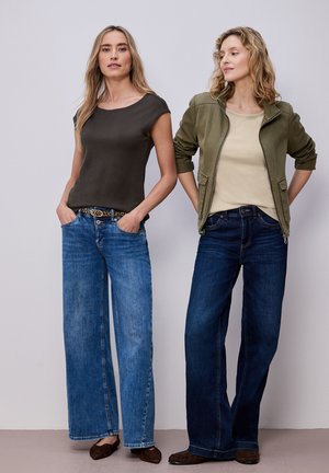 To kvinder iført vidde-benet jeans; den ene i sort top og brunt bælte, den anden i beige skjorte og grøn jakke, begge med brune sko.