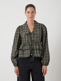 Žena nosi bluzu s kariranim uzorkom u zelenoj boji, s dugim napuhanim rukavima, velikim ovratnikom i mašnom na prednjem rufflesu, u kombinaciji s crnim hlačama visokog struka.