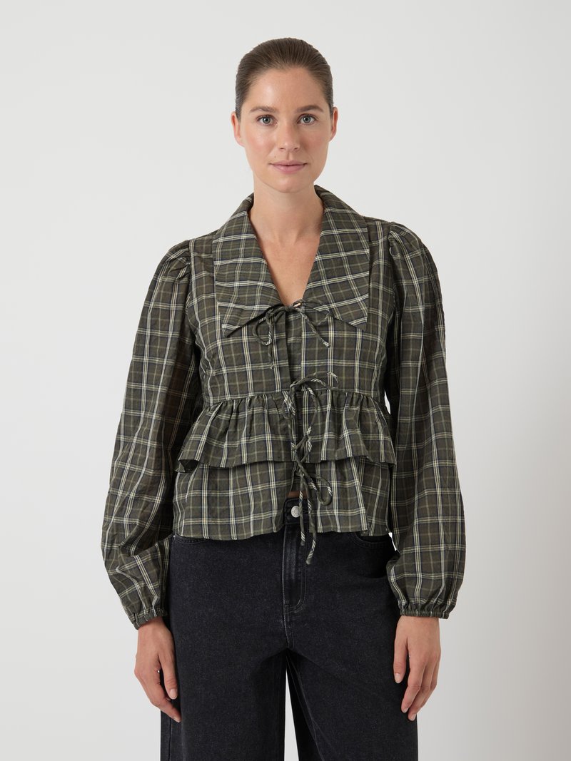 Žena nosi bluzu s kariranim uzorkom u zelenoj boji, s dugim napuhanim rukavima, velikim ovratnikom i mašnom na prednjem rufflesu, u kombinaciji s crnim hlačama visokog struka.