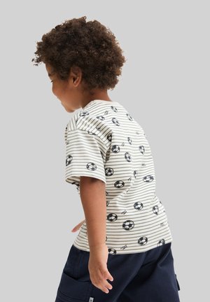 Junges Kind mit lockigem Haar, trägt ein weiß gestreiftes Hemd mit schwarzen Fußballmotiven und dunkelblaue Hose, steht seitlich vor einem grauen Hintergrund.