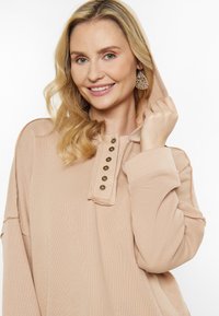 Hoodie beige con una patta di bottoni frontale, maniche lunghe e vestibilità rilassata. Il materiale ha una finitura testurizzata con una leggera costina.
