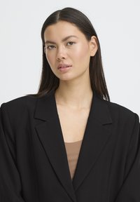 Jeune femme aux longs cheveux foncés portant un blazer noir par-dessus un haut marron, regardant légèrement sur le côté sur un fond uni.