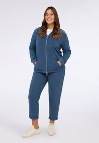 Helleblaue Kapuzenjacke mit Reißverschluss und Taschen, taillierte Bündchen und strukturierte Ärmel, kombiniert mit blauen, lässig geschnittenen Hosen und hellen Turnschuhen.