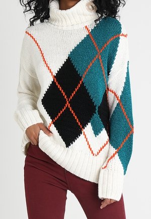 Kvinde iført en hvid rullekrave sweater med store grønblå og sorte rombemønstre med orange kant, kombineret med burgunderfarvede bukser.