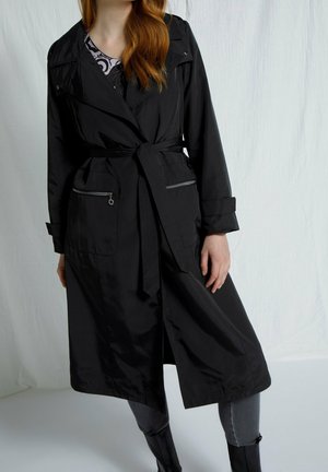 Femme portant un trench-coat noir ceinturé avec des poches zippées, un jean gris et des bottes noires sur un fond clair uni.