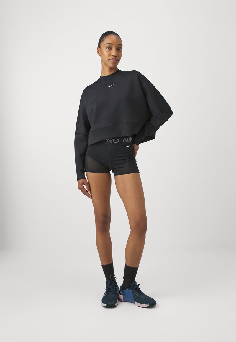 Schwarzer, kurz geschnittener Sweatshirt mit kleinem Logo, kombiniert mit schwarzen Sportshorts mit einem gebrandeten Bund. Dunkelblaue Sneakers mit pinken Akzenten.