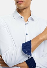 Weißes Langarmhemd mit dunkelblauen Tupfen an Manschetten und Kragen. Verfügt über blaue Knöpfe und eine glatte, leichte Textur.