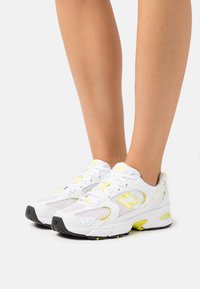 Vita atletiska skor med gula accenter, överdel i mesh och svart sula. Utrustad med snörningsdesign och förstärkt tåparti.