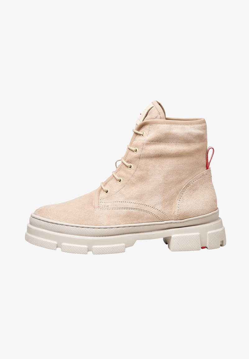 Botas de tobillo de ante beige con una suela blanca gruesa, que presentan ojales dorados, una tira roja para tirar y acentos laterales texturizados.