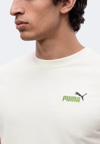 Νέος άνδρας που φοράει λευκό t-shirt Puma με πράσινο λογότυπο και μαύρο εικονίδιο Puma στο αριστερό μέρος του στήθους, κοιτάζοντας προς τα αριστερά σε κοντινή λήψη.