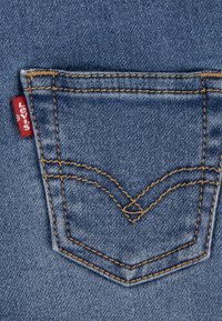 Denimstoff baklomme med oransje søm, som har et karakteristisk avrundet design og en rød merkelapp sydd på siden.