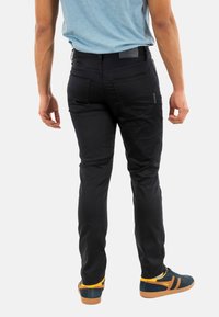 Pantalons slim noirs au toucher lisse et avec des détails subtils sur les poches, associés à des baskets vertes et jaunes, vus de dos.
