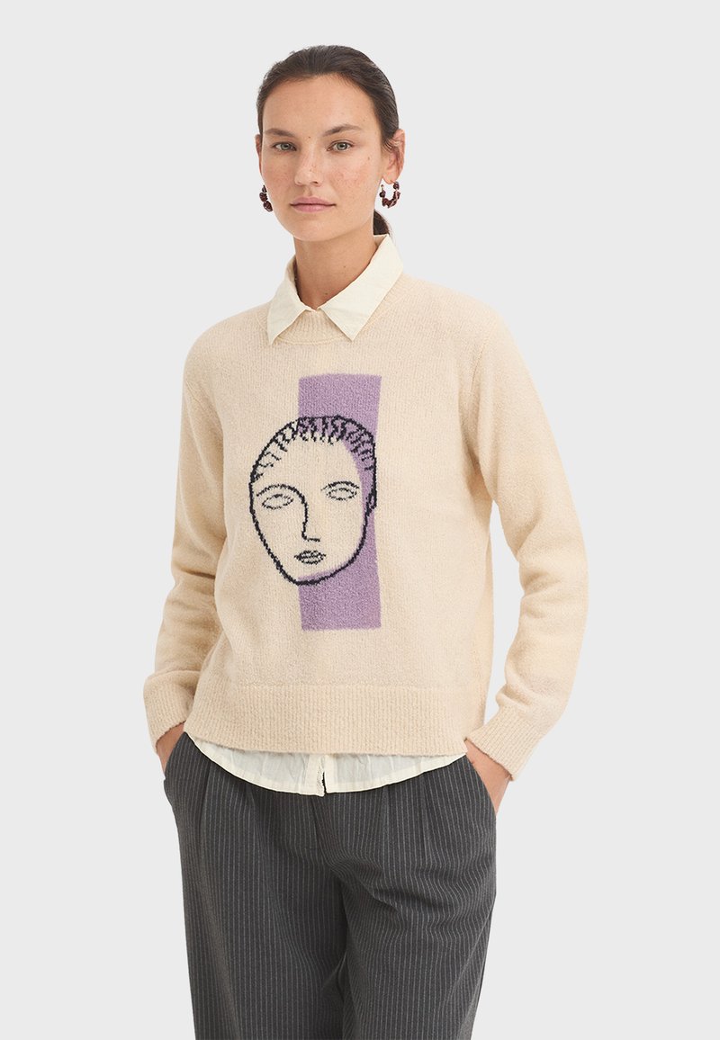 Beige sweater with front purple rectangle and black outline of a face. Knitted texture, long sleeves, paired with a white collared shirt and striped pants.