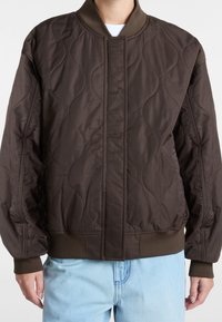 Brązowa pikowana kurtka bomberka z prążkowanym kołnierzem, posiadająca teksturowane wzory i elastyczne mankiety. W zestawie z jasnoniebieskimi dżinsami.