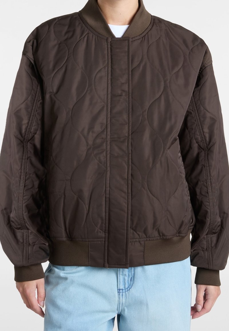 Brązowa pikowana kurtka bomberka z prążkowanym kołnierzem, posiadająca teksturowane wzory i elastyczne mankiety. W zestawie z jasnoniebieskimi dżinsami.