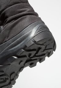 Bota de senderismo negra con parte superior de tela texturizada y una suela de goma resistente con tacos profundos para tracción. Primer plano del área del talón.