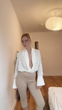 Bela bluza s globokim V-izrezom in dolgimi razširjenimi rokavi, v kombinaciji z bež leginskimi hlačami visoke pasu. Gladek material in nevtralna barvna paleta.