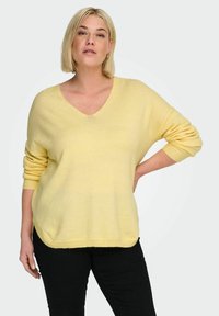 Pull-over oversize à col en V en maille jaune douce. Présente des manches retroussées et un ourlet côtelé. Porté avec un pantalon noir.