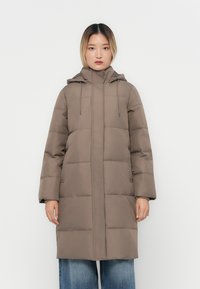Manteau poivre marron avec capuche, doté d'une fermeture éclair, de cordons élastiques et de grandes sections matelassées. Le modèle le porte sur un jean ample bleu.