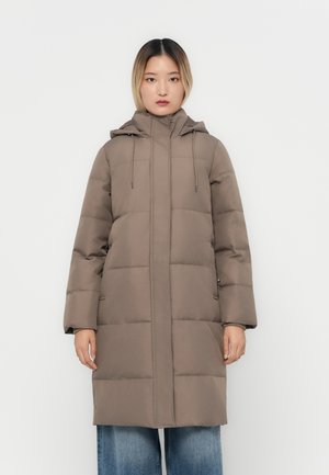 Manteau poivre marron avec capuche, doté d'une fermeture éclair, de cordons élastiques et de grandes sections matelassées. Le modèle le porte sur un jean ample bleu.