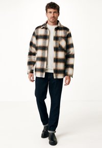Camicia di flanella a quadri in tonalità crema, marrone e nera, con chiusura a bottoni, taschino sul petto e vestibilità comoda. Indossata con pantaloni di jeans scuri e stivali.