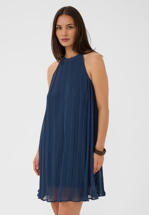 Femme aux longs cheveux foncés portant une robe plissée bleu marine sans manches et un bracelet sombre au poignet droit, debout devant un fond uni.