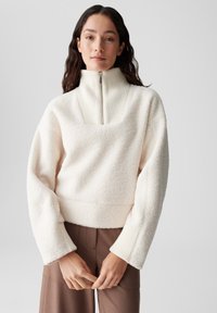 Fleece-Pullover in Off-White mit geripptem Stehkragen. Hat einen eckigen Ausschnitt und weite Ärmel, kombiniert mit braunen Hosen.
