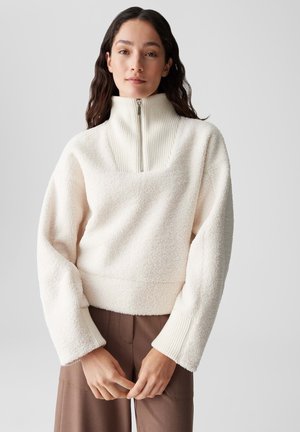 Fleece-Pullover in Off-White mit geripptem Stehkragen. Hat einen eckigen Ausschnitt und weite Ärmel, kombiniert mit braunen Hosen.