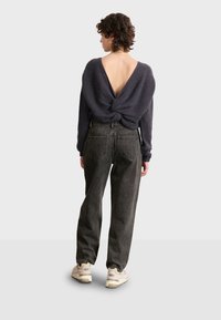 Suéter de punto gris oscuro con un profundo escote en la espalda, detalle torcido en la parte baja de la espalda, combinado con jeans negros de cintura alta y deportivas claras.