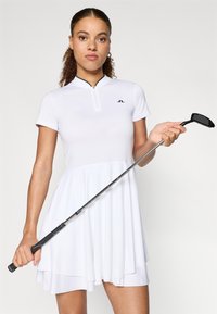 Biała sukienka golfowa wykonana z gładkiej tkaniny, z kołnierzykiem na zamek błyskawiczny i rozkloszowanym dołem. Modelka trzyma kij golfowy z czarną główką.