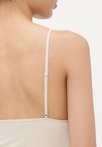 Close-up van de bovenrug en schouder van een persoon die een beige spaghettibandtop met een verstelbare band draagt, tegen een effen witte achtergrond.