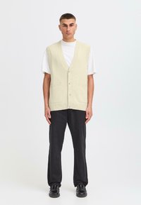 Un uomo è in piedi su uno sfondo neutro, indossando un gilet di maglione crema sopra una maglietta bianca e pantaloni scuri, con scarpe nere.