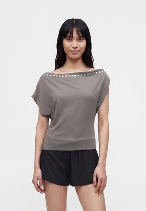 Kvinde med langt mørkt hår iført grå off-shoulder top med sølv nitter og sorte shorts, står foran en ensfarvet lys baggrund.