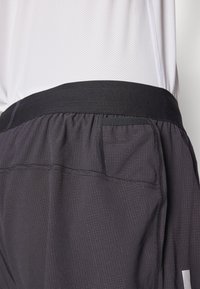 Schwarze Sportshorts aus strukturiertem Stoff, mit elastischem Bund und seitlichem Taschendetail. Ein dezentes Karomuster ist auf dem Material sichtbar.