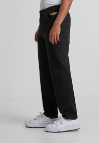 Jeans en denim noir avec une coupe droite, dotés de poches avant et d'un patch de logo coloré au niveau de la taille. Portés avec des baskets blanches.