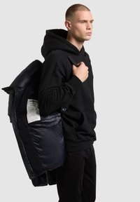 Čierna mikina s kapucňou a prednou kapsou, doplnená o námornícku prešívanú bundu držanú na ramene. Bunda má viditeľný zips a štítok vo vnútri.