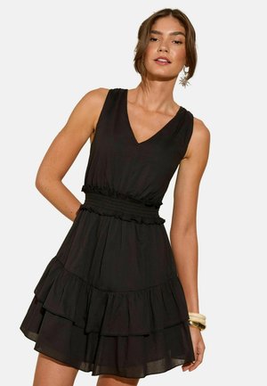 Schwarzes Mini-Kleid mit V-Ausschnitt, ärmellosem Design, gesmockter Taille und gestuftem Rüschensaum. Hergestellt aus leichtem Stoff.